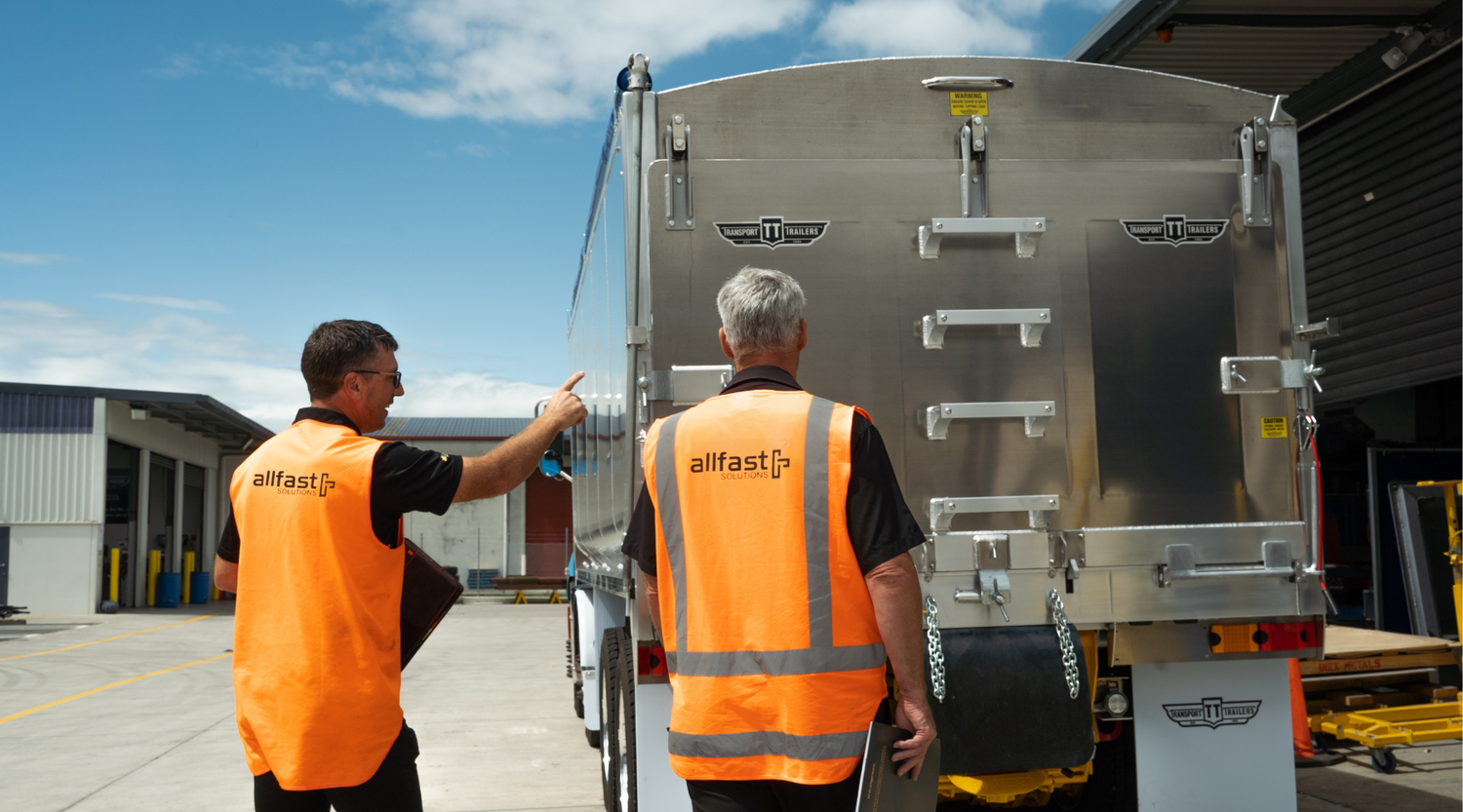 Two workers in orange safety vests inspecting a large metallic trailer outdoors.